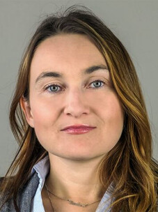 Porträtfoto von Ewelina M. Hankiewicz mit offenen Haaren und dezentem Lächeln vor grauem Hintergrund.